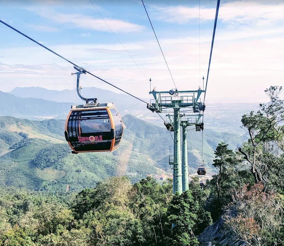 Một cabin cáp treo của Sun World Ba Na Hills trên đường cáp, cột trụ màu xanh, bên dưới là rừng và núi