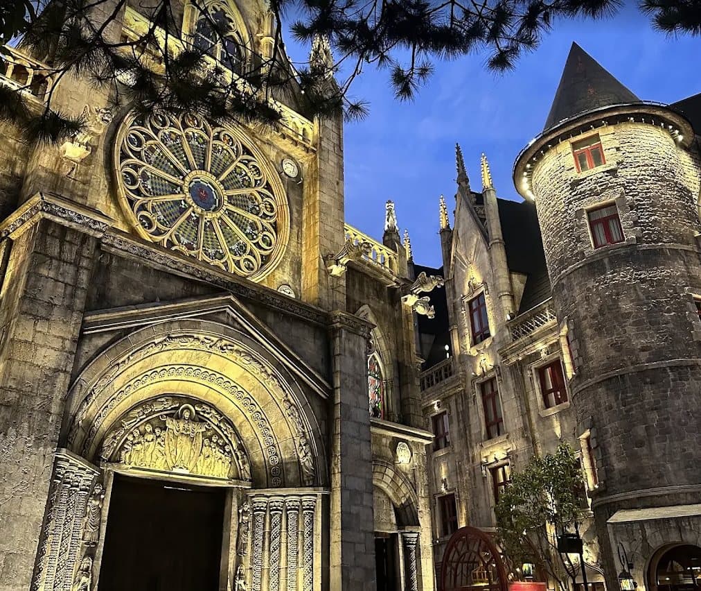 Mặt tiền nhà thờ có cửa sổ hoa hồng và cửa vòm chạm khắc trong Làng Pháp tại Sun World Ba Na Hills, lên đèn lúc chạng vạng, bên cạnh một tháp tròn