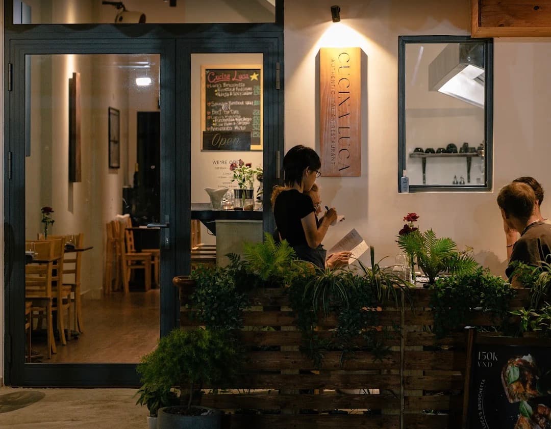 Cucina Luca, mặt trước nhà hàng vào buổi tối, với bảng tên, bảng phấn và khách ngồi bàn bên ngoài