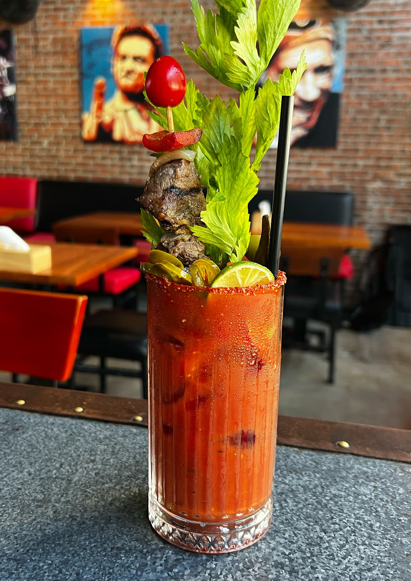 A bloody mary with a skewer of grilled beef in it on the bar at Dirty Fingers, with brick walls, red booths and painted portraits behind