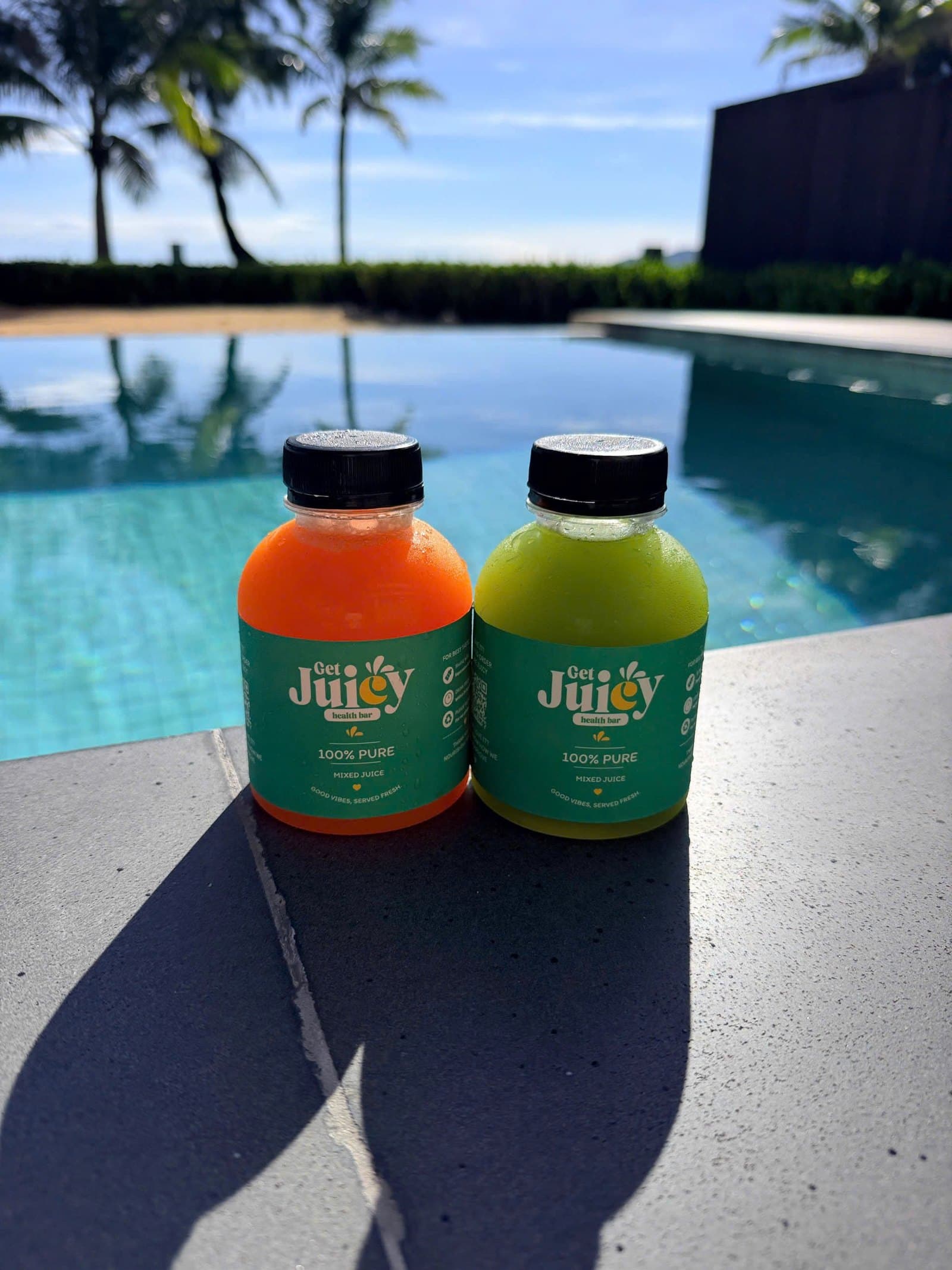 Two bottles of GetJuicy Health Bar mixed juice, one orange and one green, on the edge of a pool