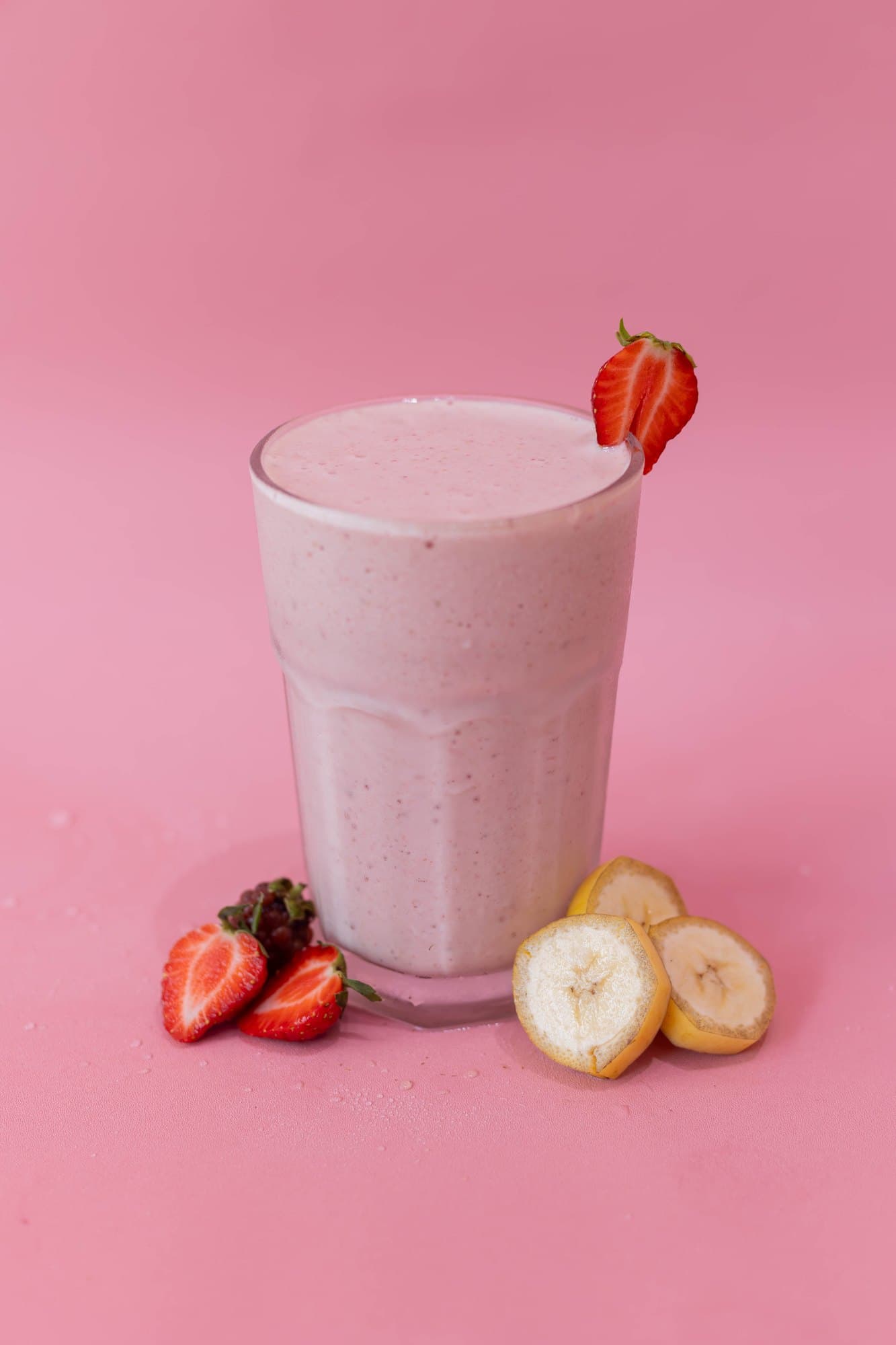 A pink GetJuicy Health Bar smoothie in a glass with a strawberry on the rim, beside strawberries and banana