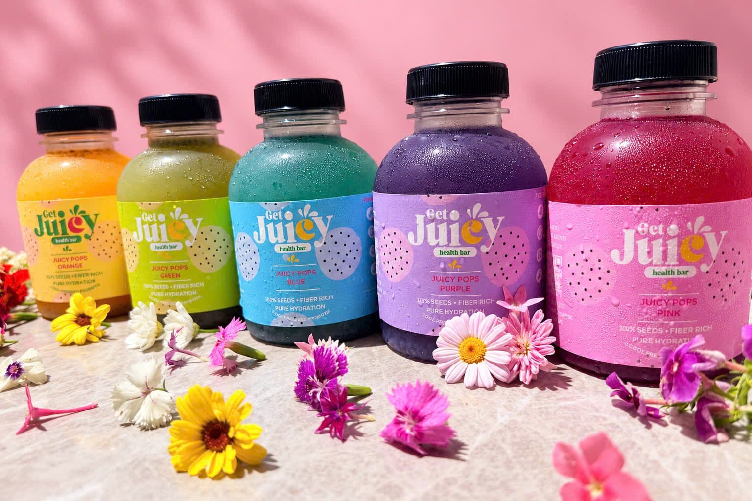 Five Juicy Pops bottles from GetJuicy Health Bar, yellow, green, blue, purple and pink, with flowers on a pink background