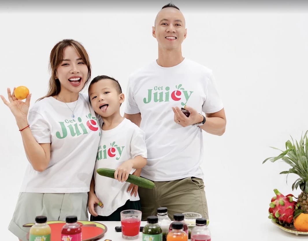 A mother, her son and a father in GetJuicy Health Bar T-shirts at a table of GetJuicy Health Bar bottles; the mother holds an orange and the son a cucumber