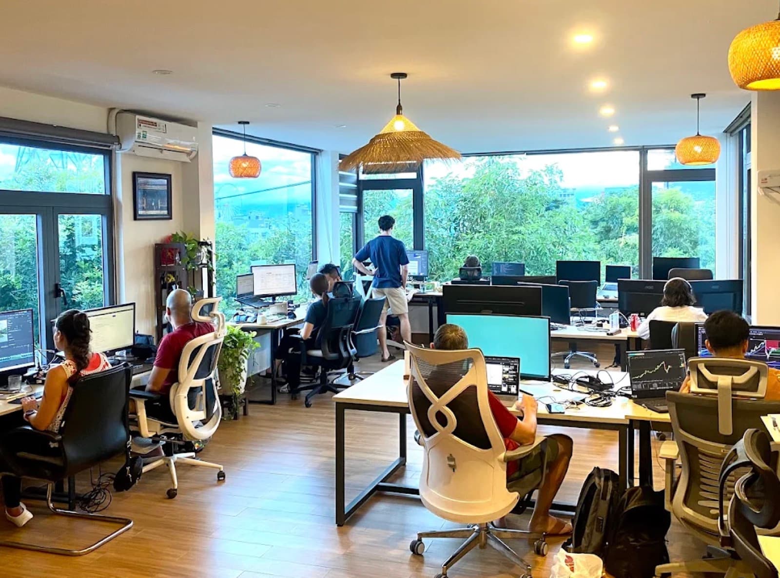 The working floor at Hana's Coworking, desks and monitors beside windows onto the trees in Da Nang