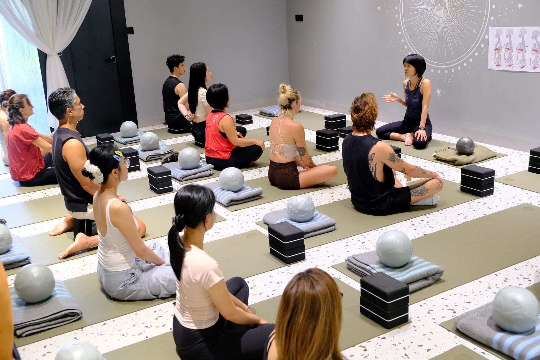 Cả lớp ngồi trên thảm xanh ở Heart Studio by Chan, hướng về một người phụ nữ phía trước, cạnh mỗi thảm có bóng, chăn gấp và gạch yoga