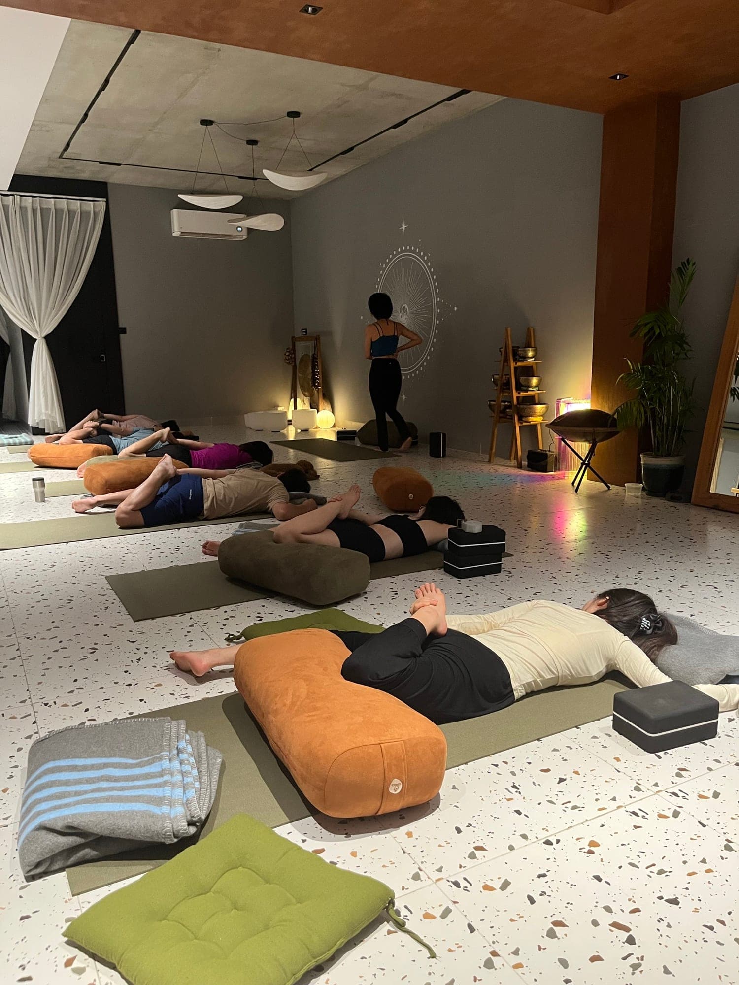 Học viên nằm trên gối bolster vào buổi tối ở Heart Studio by Chan, một người phụ nữ đứng cạnh hình mặt trăng trên tường