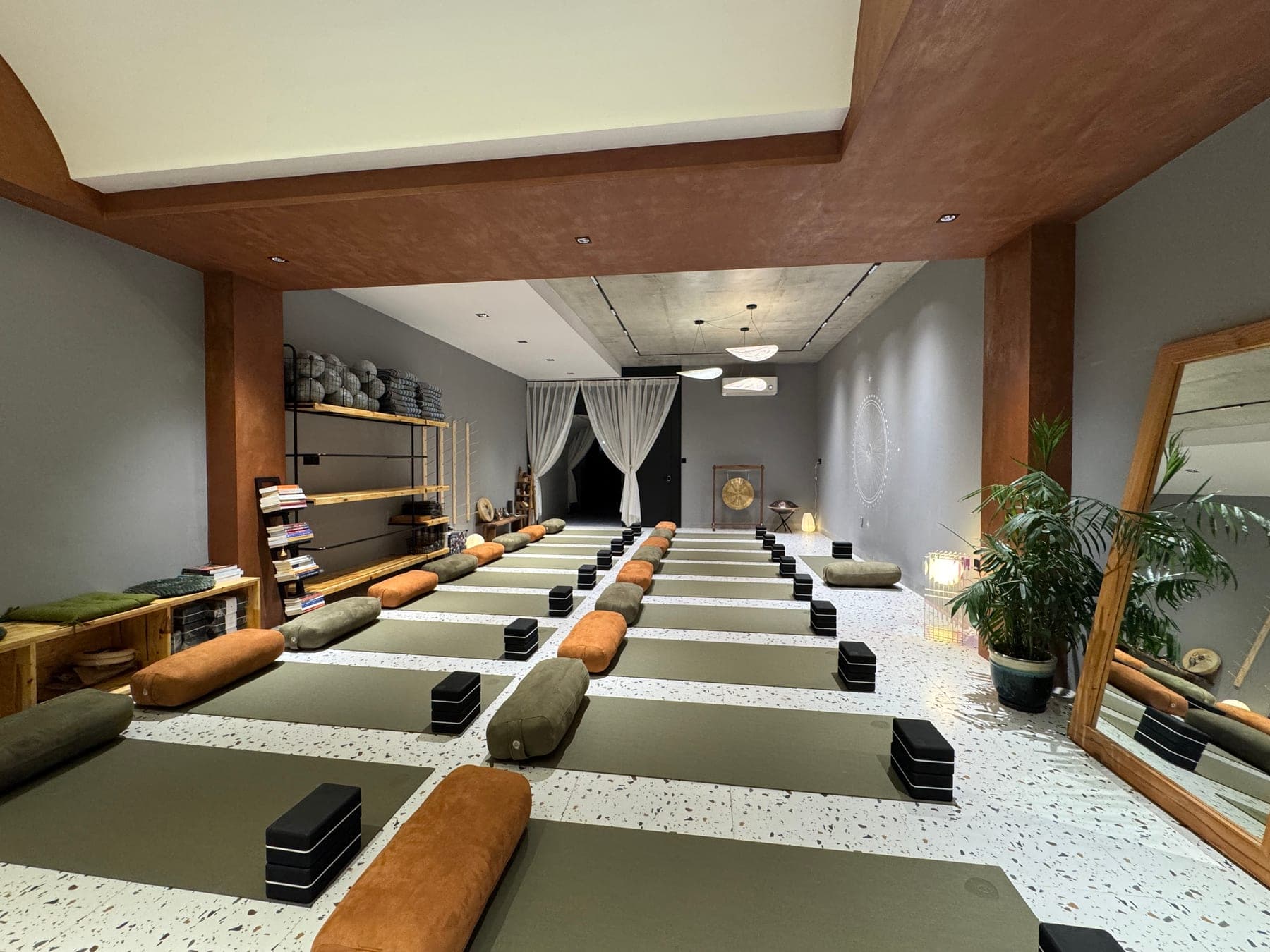 Phòng tập trống ở Heart Studio by Chan: những hàng thảm xanh, gối bolster cam và xanh, gạch yoga đen trên sàn đá mài