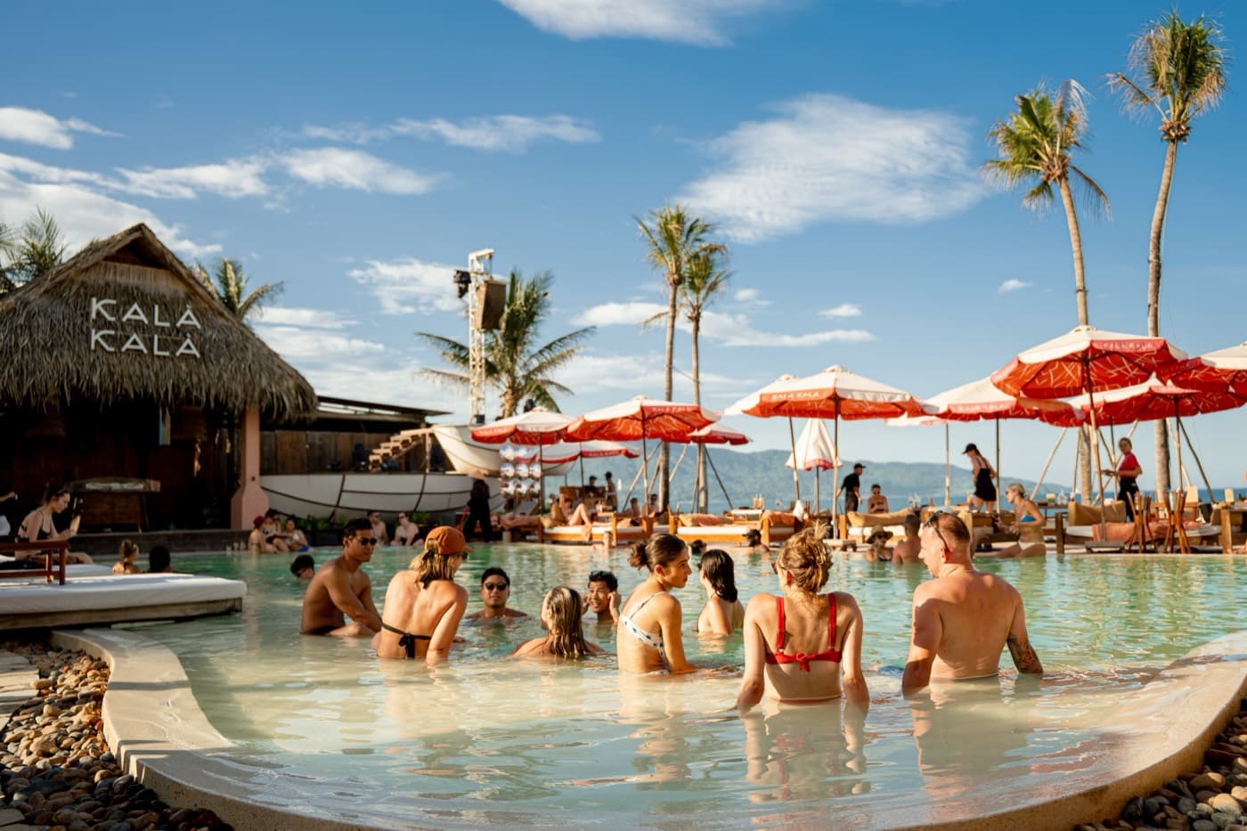Kalà Kalà Beach Club, people sitting in the pool in the sun, a thatched hut with the Kala Kala name, red and white umbrellas and palm trees