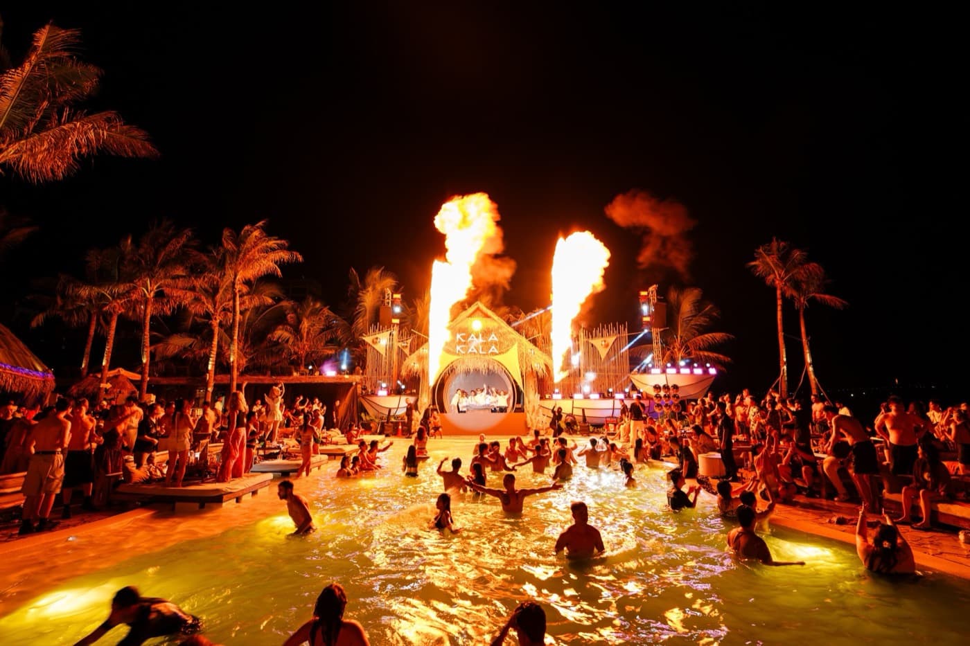Kalà Kalà Beach Club, the DJ booth under the Kala Kala sign at night, with jets of fire and a crowd in the lit pool