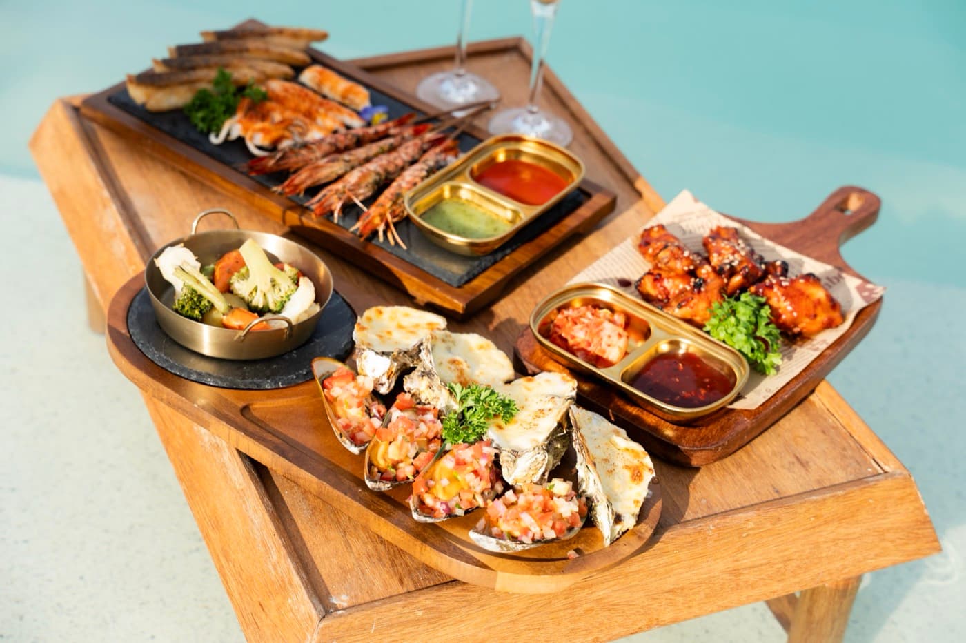 Kalà Kalà Beach Club, grilled prawns, oysters with toppings, glazed wings and roasted vegetables on wooden boards by the pool