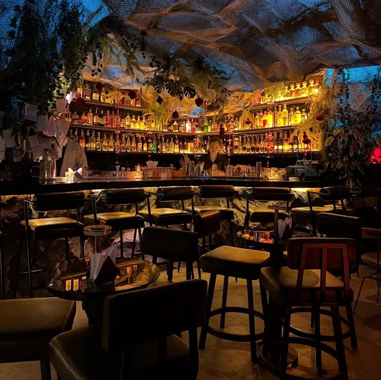 The bar at Ket High Cocktail Bar with no guests: stools, lit shelves of bottles and draped mesh above