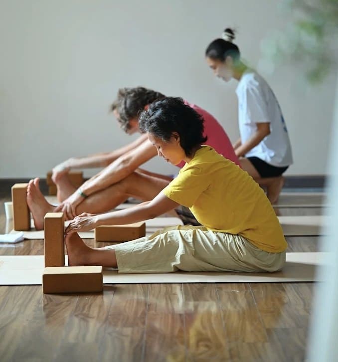 Ba người cúi gập về phía trước trên thảm tập tại Mandir Danang, có gạch yoga bằng gỗ bần