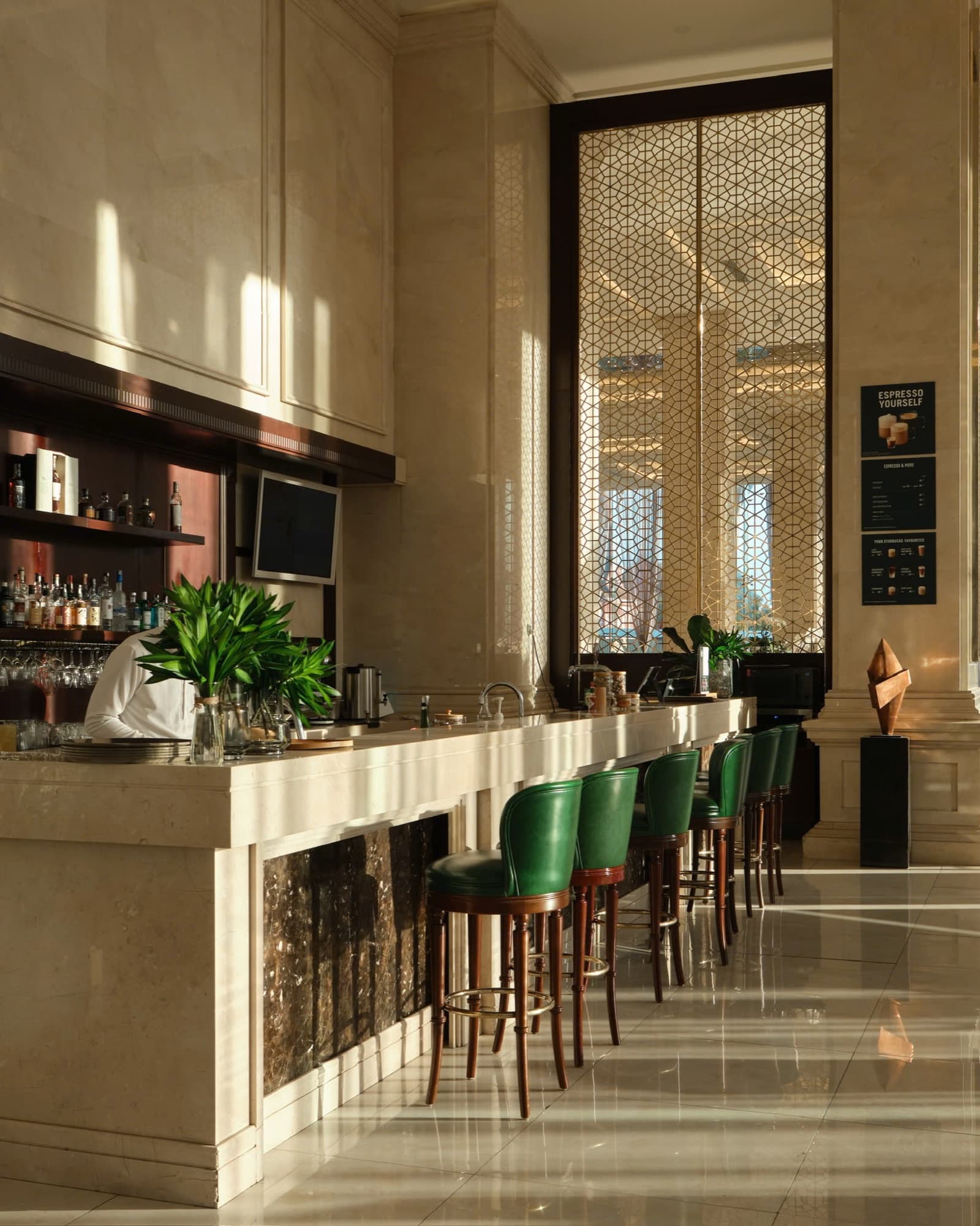 Quầy bar tại Meliá Vinpearl Danang Riverfront trong nắng xiên: hàng ghế da xanh lá dọc quầy đá cẩm thạch, cây xanh và tấm bình phong hoa văn lưới màu vàng kim