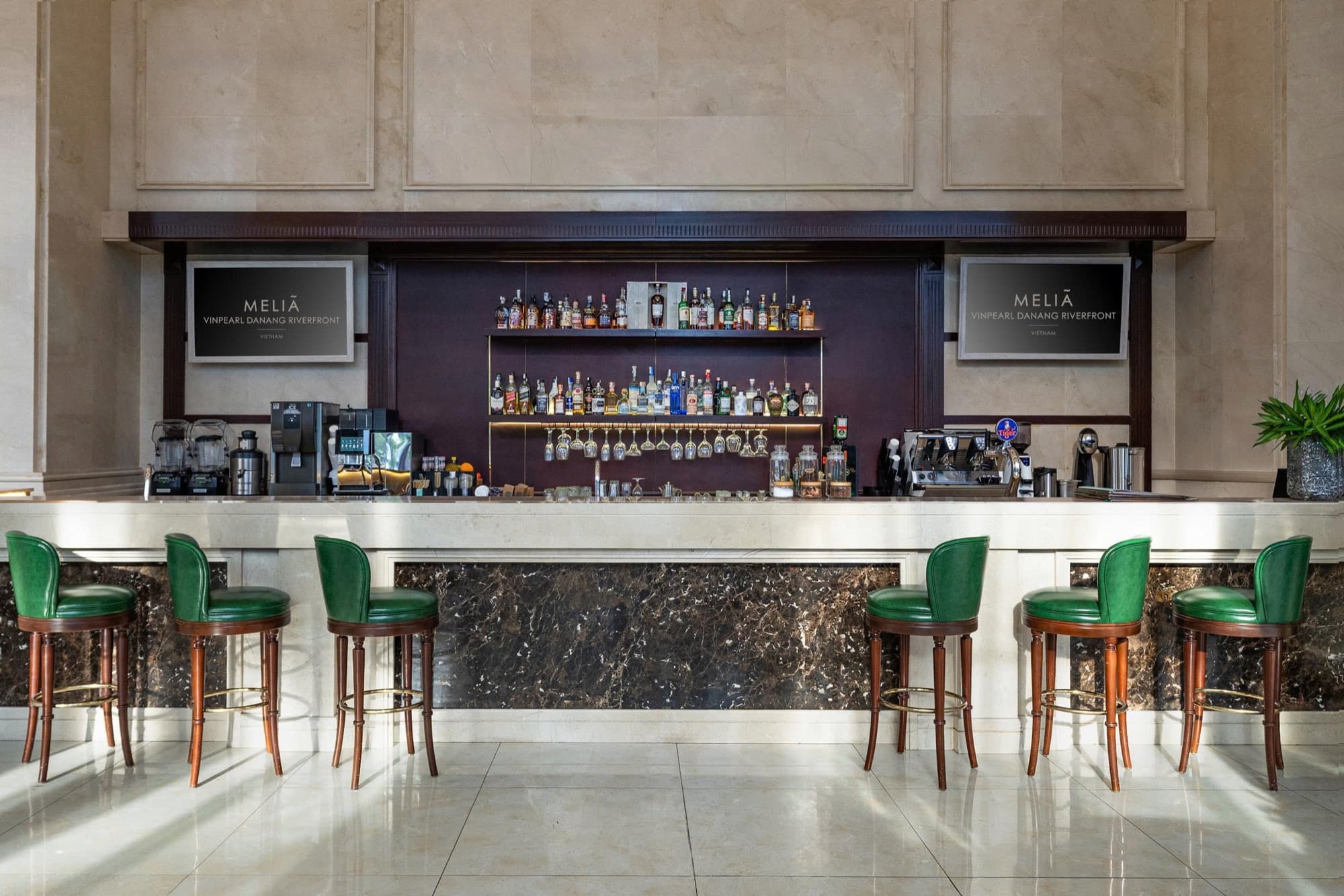 Quầy bar tại Meliá Vinpearl Danang Riverfront: ghế da xanh lá bên quầy đá cẩm thạch, chai rượu trên kệ có đèn, và hai màn hình ghi Meliá Vinpearl Danang Riverfront