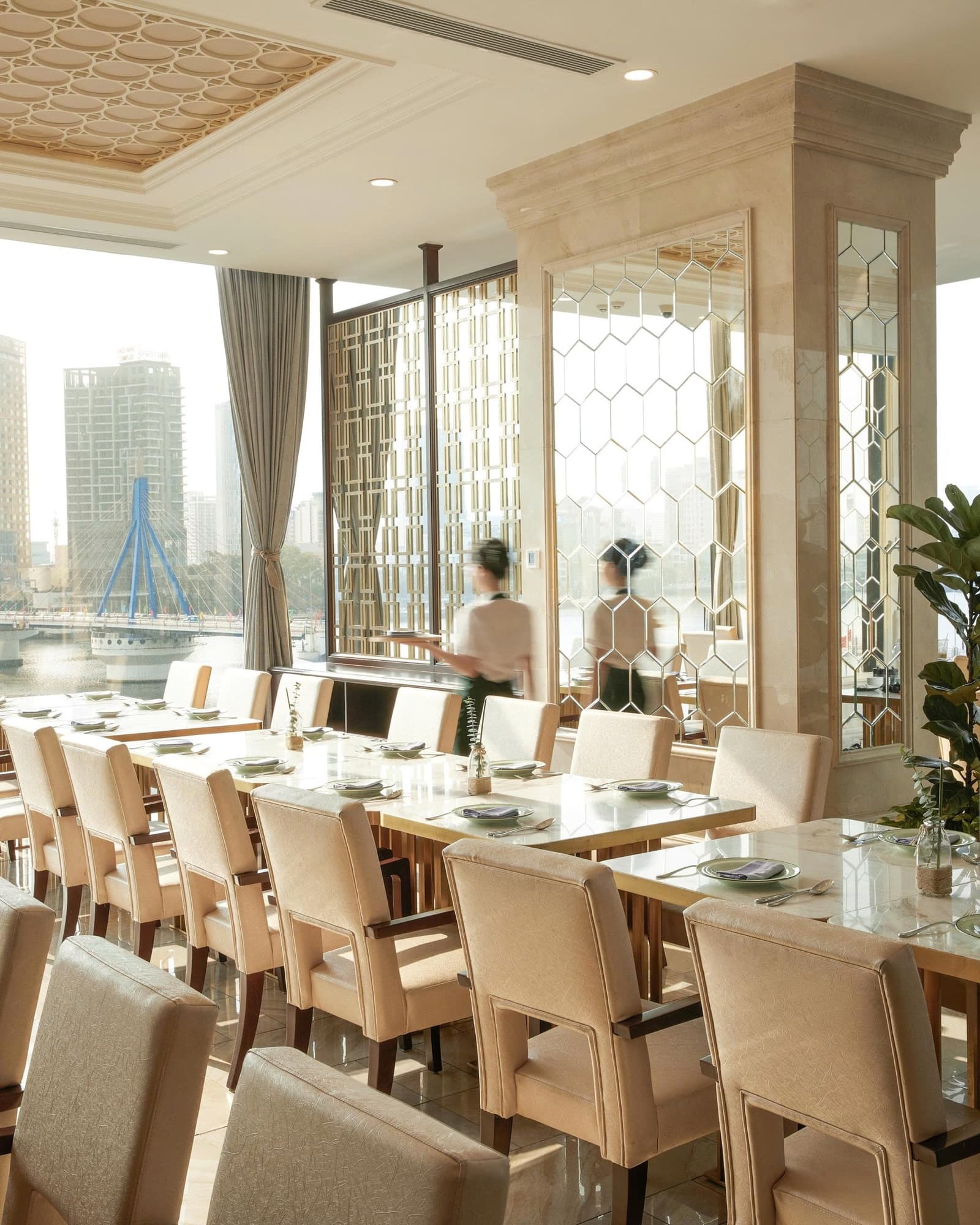 Nhà hàng Han River 1 tại Meliá Vinpearl Danang Riverfront ban ngày: bàn dài đã bày đĩa, một nhân viên bưng khay và cầu sông Hàn ngoài cửa sổ