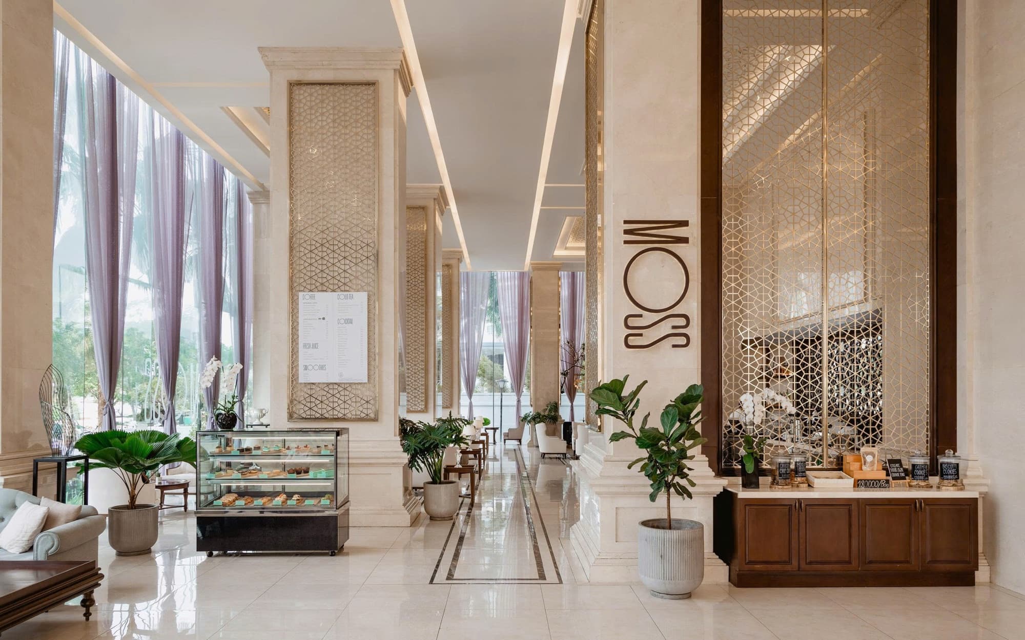 Moss Lounge tại Meliá Vinpearl Danang Riverfront: chữ MOSS trên cột đá cẩm thạch, tủ kính bánh ngọt, hũ bánh quy, bảng thực đơn và cửa sổ cao có rèm