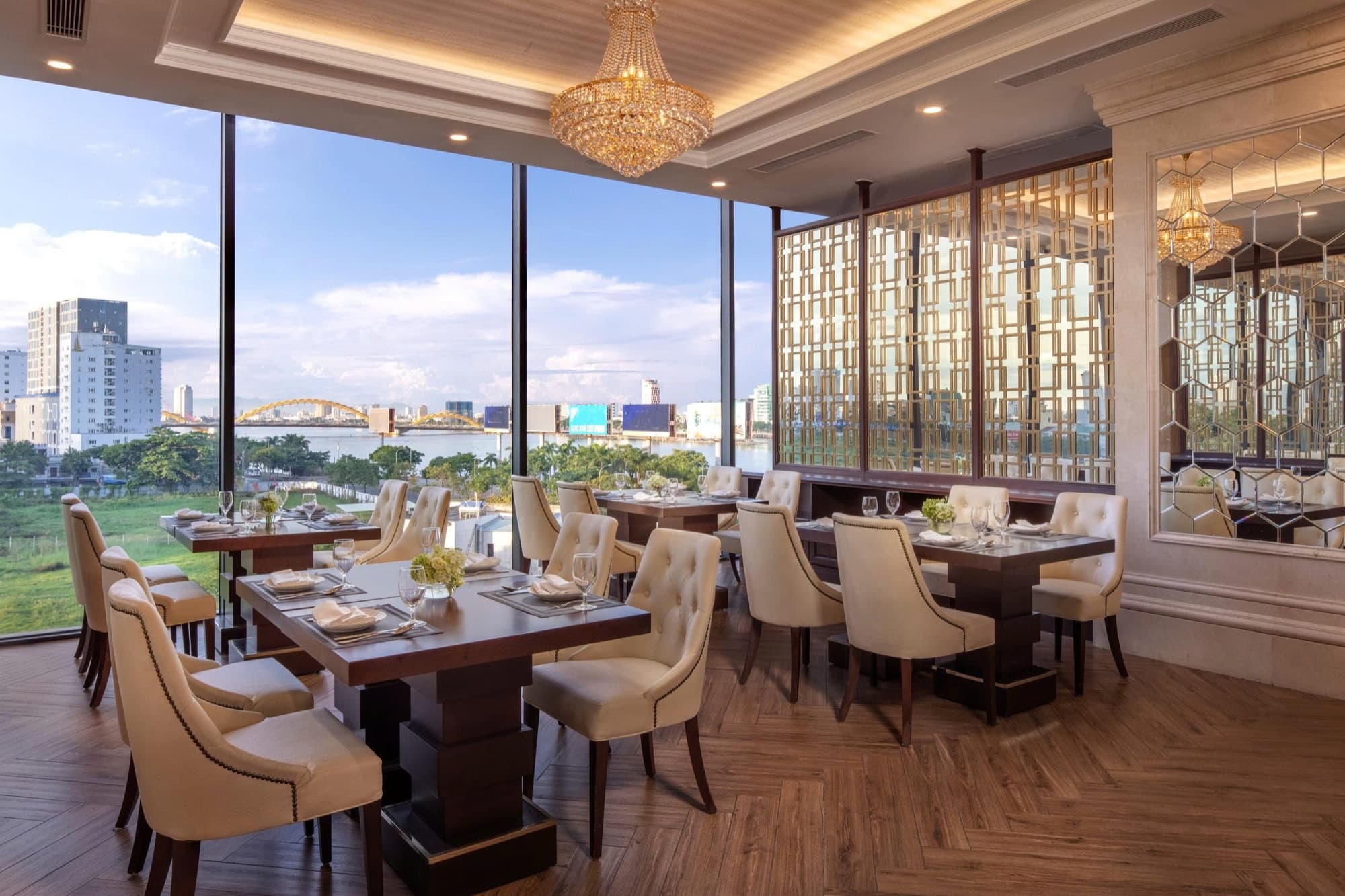 Một phòng ăn tại Meliá Vinpearl Danang Riverfront ban ngày: ghế màu kem, ly rượu vang trên bàn gỗ tối màu, đèn chùm pha lê và cầu Rồng ở xa sau vách kính cao từ sàn đến trần