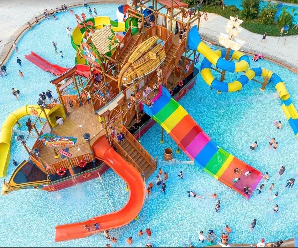 Mikazuki Water Park 365, tàu cướp biển ngoài trời nhìn từ trên cao, có đường trượt bảy màu, đường trượt đỏ và ống trượt xanh dương và vàng, giữa hồ bơi đông người