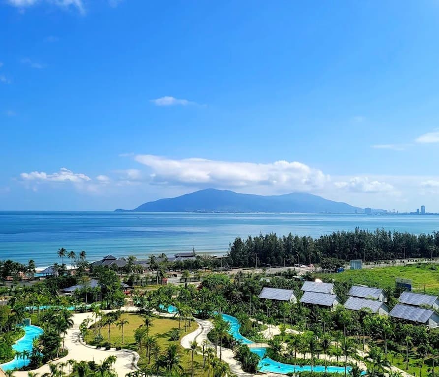 Mikazuki Water Park 365, the outdoor pools and white paths among palm trees, seen from above, with rows of villa roofs, the sea and a mountain behind
