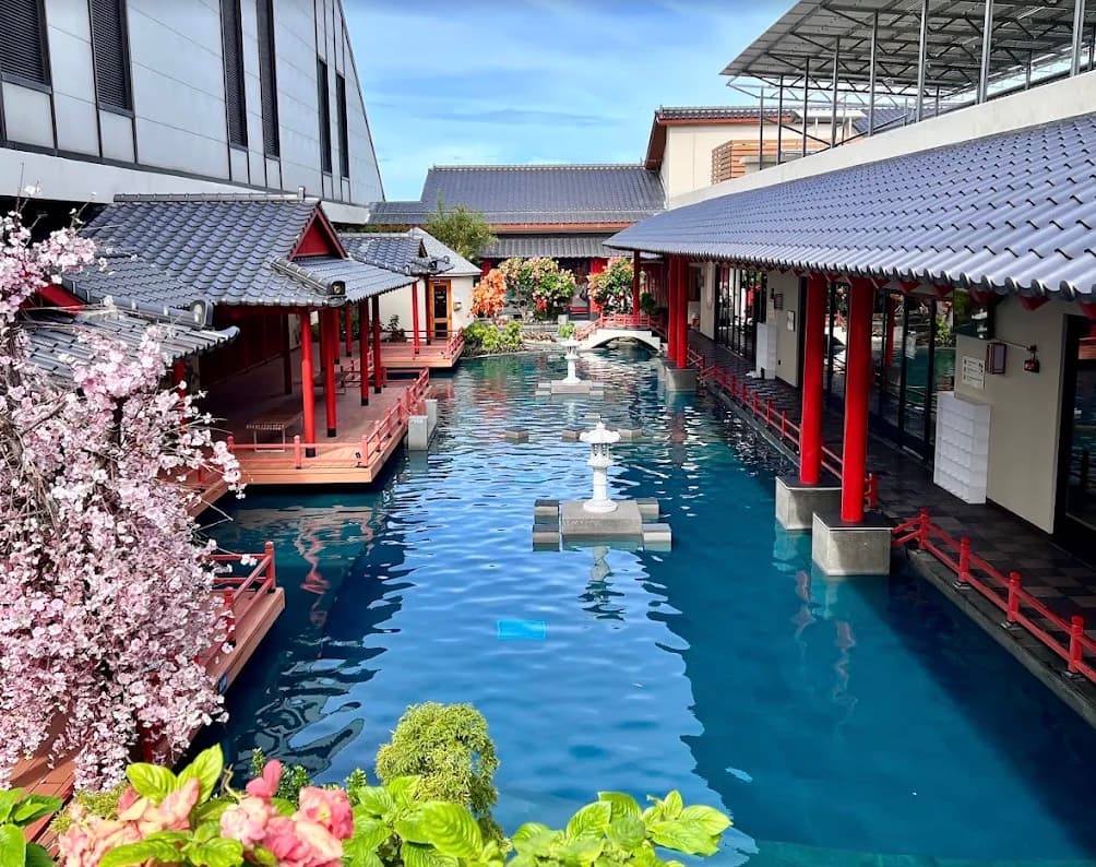 Mikazuki Water Park 365, khoảng sân kiểu Nhật có cột đỏ, mái ngói xám và những chiếc đèn đá đứng giữa hồ nước xanh, phía trước có hoa màu hồng