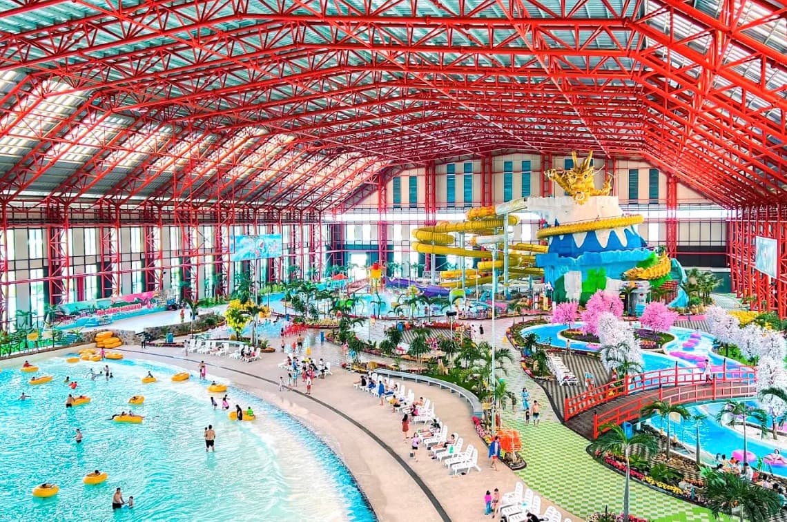 Mikazuki Water Park 365, khu trong nhà dưới khung mái thép màu đỏ, có bể tạo sóng với phao vàng, tháp đường trượt với con rồng vàng trên đỉnh, những cây hoa màu hồng và băng rôn ĐÀ NẴNG MIKAZUKI trên bức tường phía xa