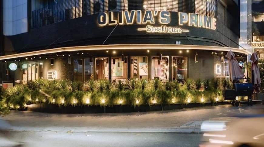 The corner of Indochina Mall at dusk, with the lit Olivia's Prime Steakhouse sign and plants along the pavement