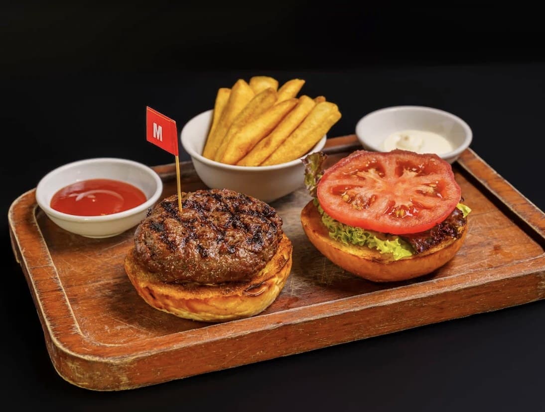 A hamburger at Olivia's Prime Steakhouse on a wooden tray, with fries, ketchup, a white sauce and a small red flag marked M