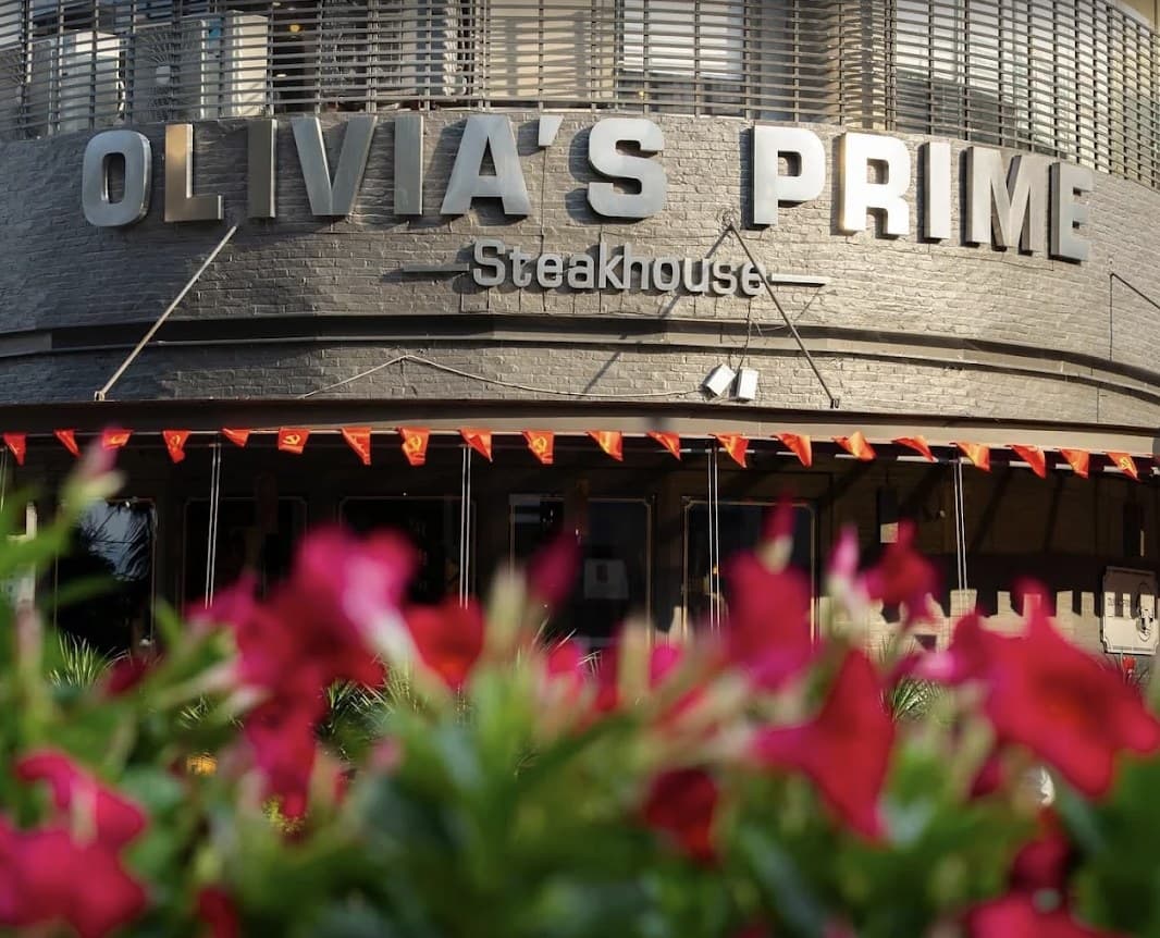 The Olivia's Prime Steakhouse sign over the front of the restaurant, with red pennant flags and red flowers in front