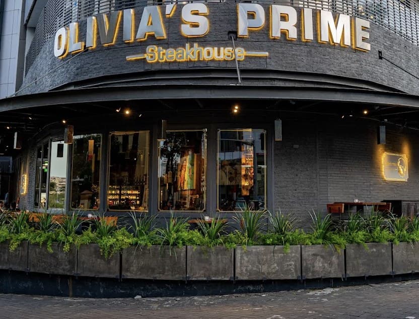 The front of Olivia's Prime Steakhouse in the evening: the lit name on the corner, tall windows, and green plants in long planters