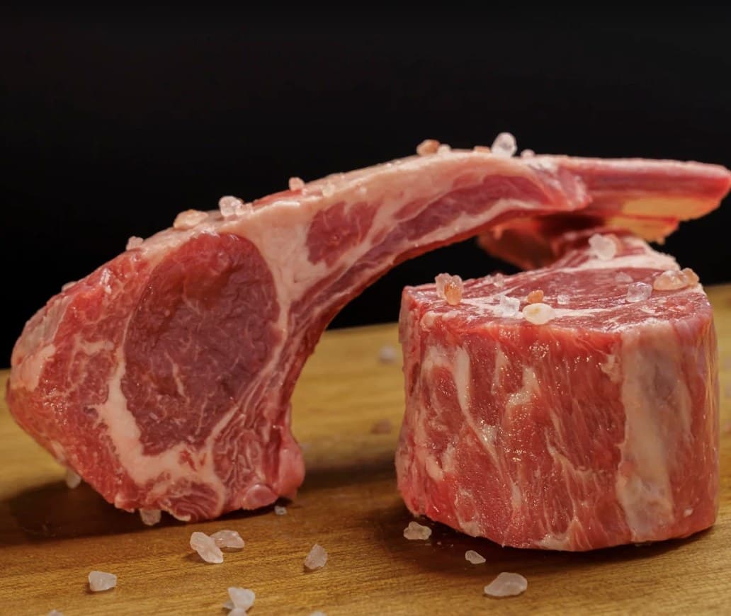 Two raw cuts of beef with coarse salt on a wooden board at Olivia's Prime Steakhouse, one on a long bone