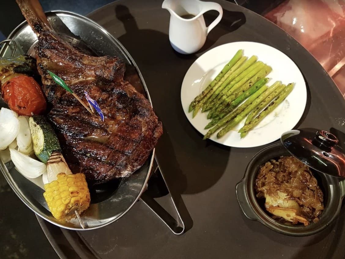 A long bone steak at Olivia's Prime Steakhouse on a metal plate with grilled vegetables and corn, with asparagus and a covered pot beside it
