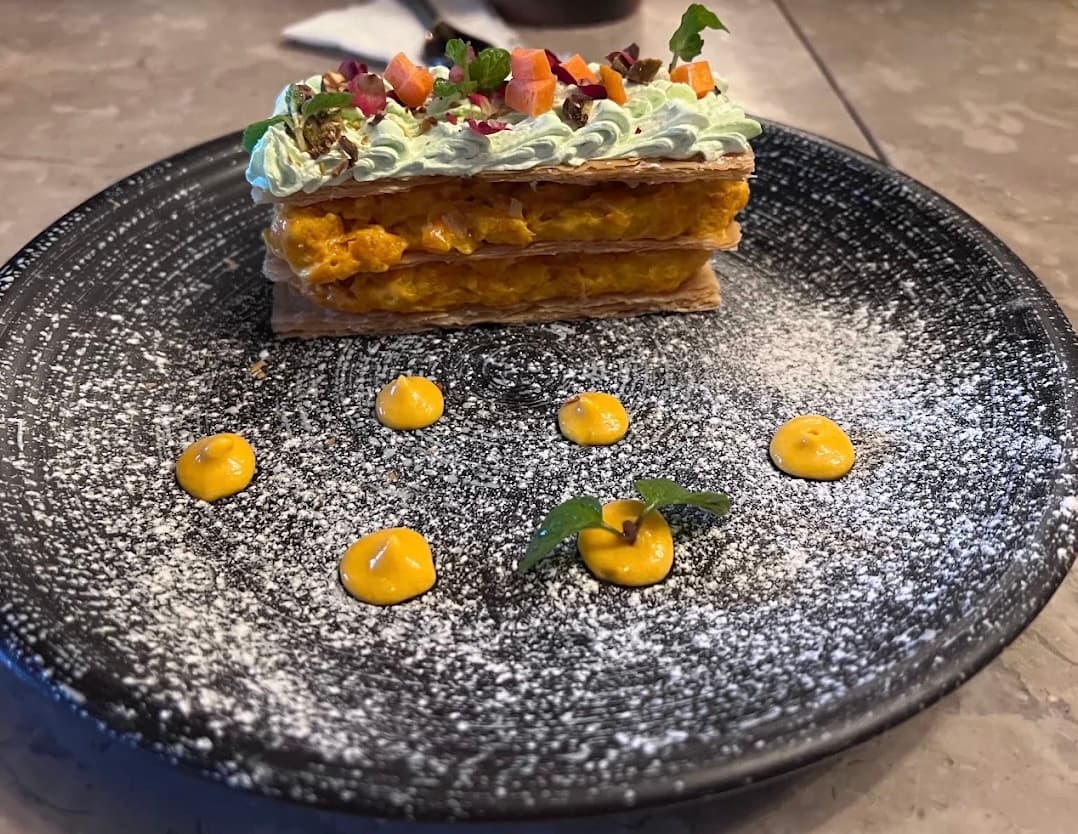 A layered pastry dessert with saffron cream and pistachio on a dark plate dusted with sugar at RANG