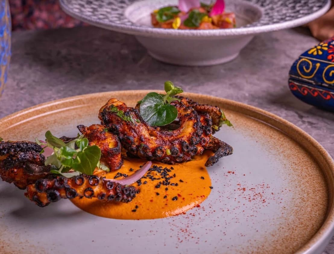 Charred tandoori octopus on a smear of almond romesco, scattered with black sesame and herbs, on a stone plate at RANG