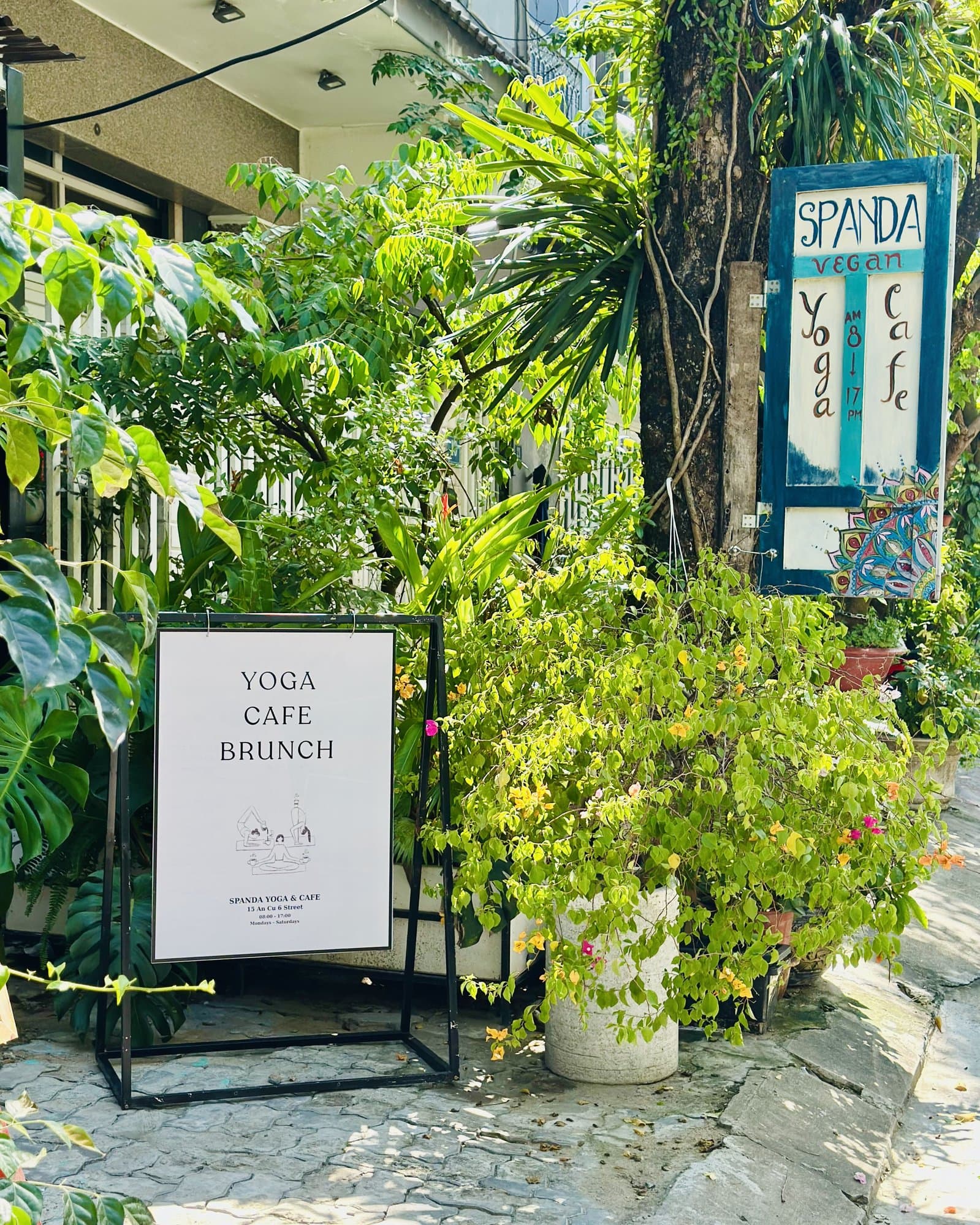 Lối vào Spanda Yoga: tấm biển vẽ tay chữ Spanda treo trên cây và tấm bảng ghi Yoga, Cafe, Brunch giữa cây xanh