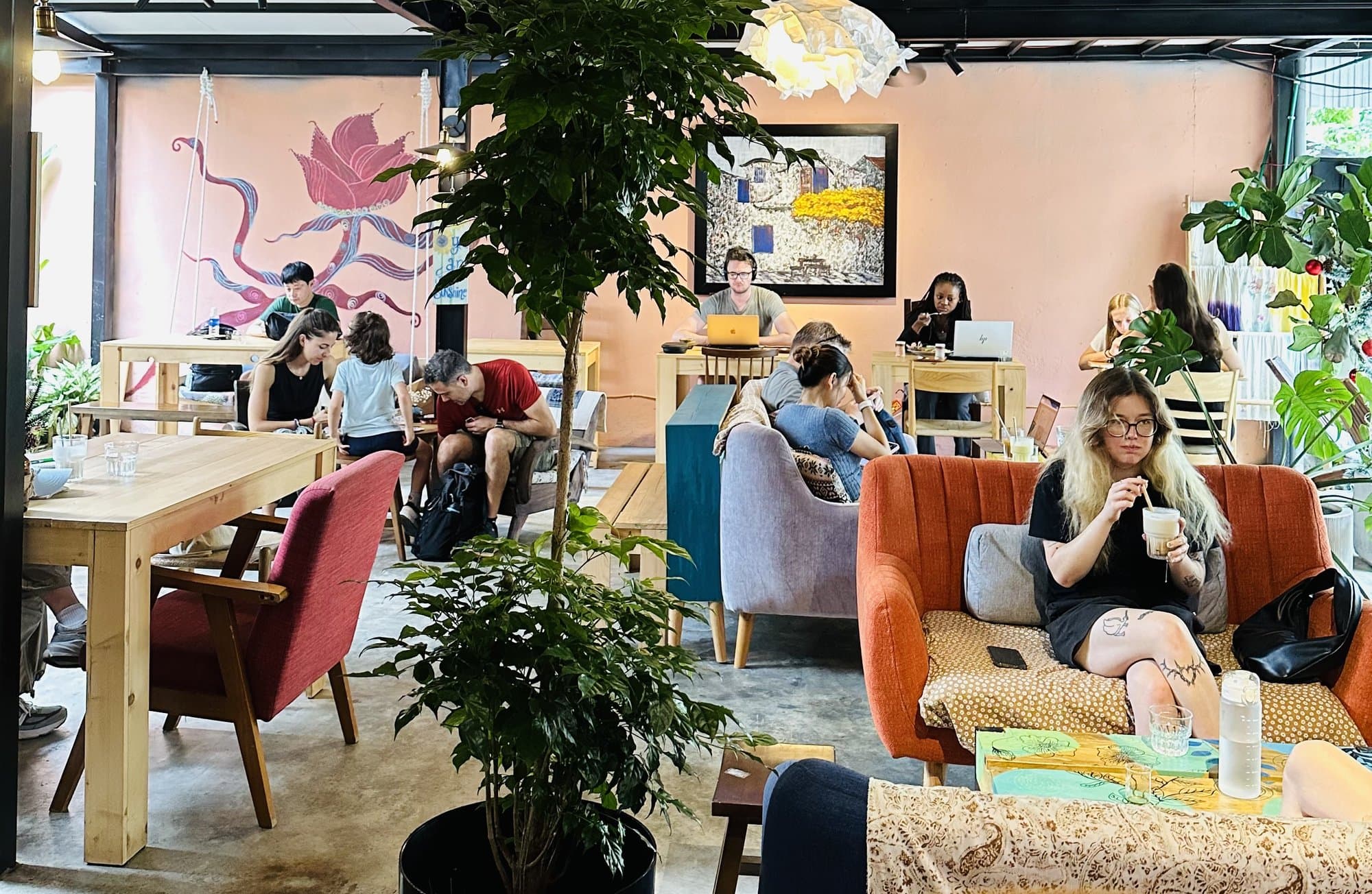 Quán cà phê tại Spanda Yoga: bàn gỗ, ghế sofa cam, cây xanh, tranh treo trên tường màu hồng đào và khách ngồi làm việc với laptop
