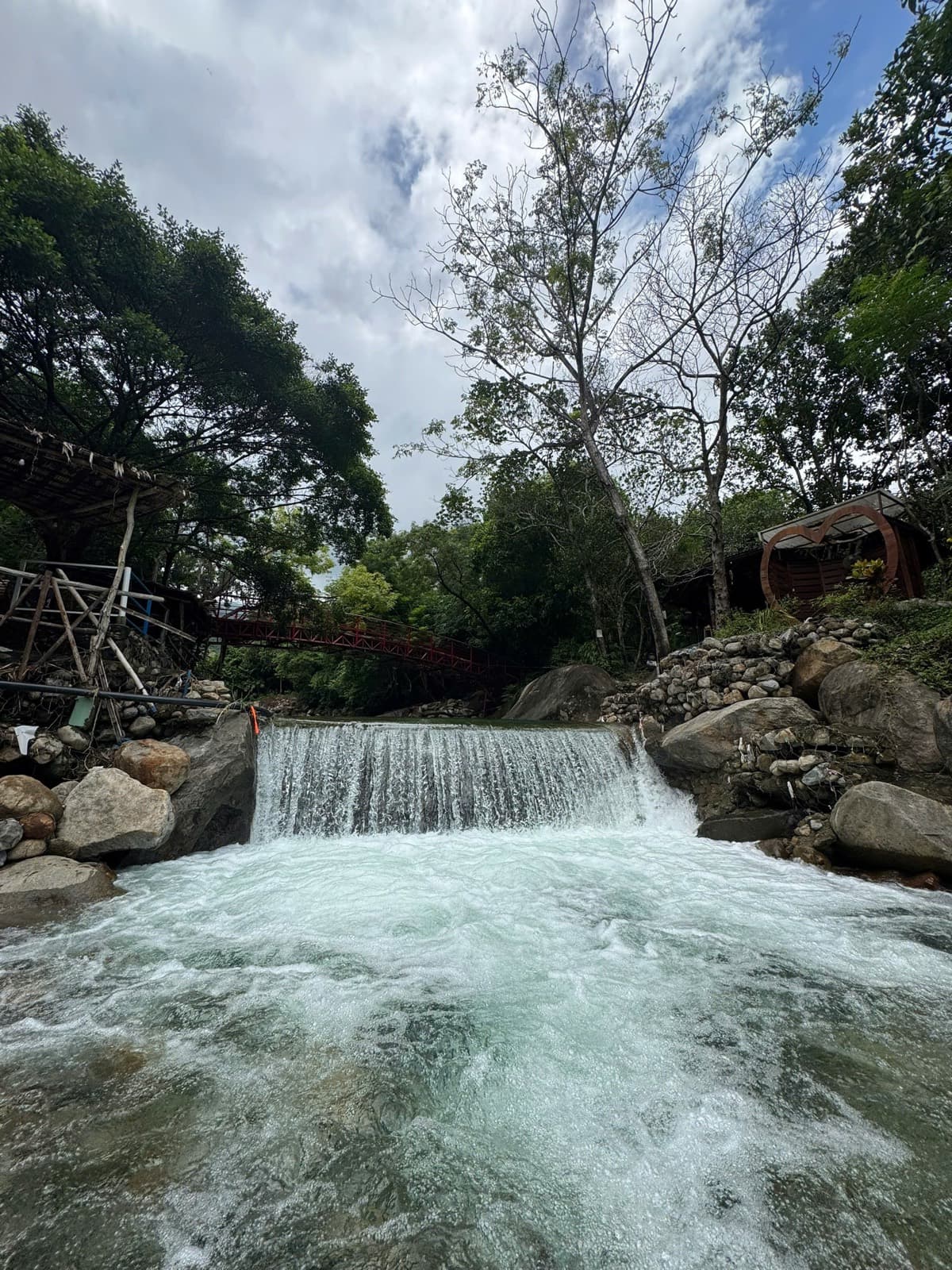 Suối Lương Hải Vân Park, water pouring over a low weir in the stream, with a red bridge and wooden huts behind