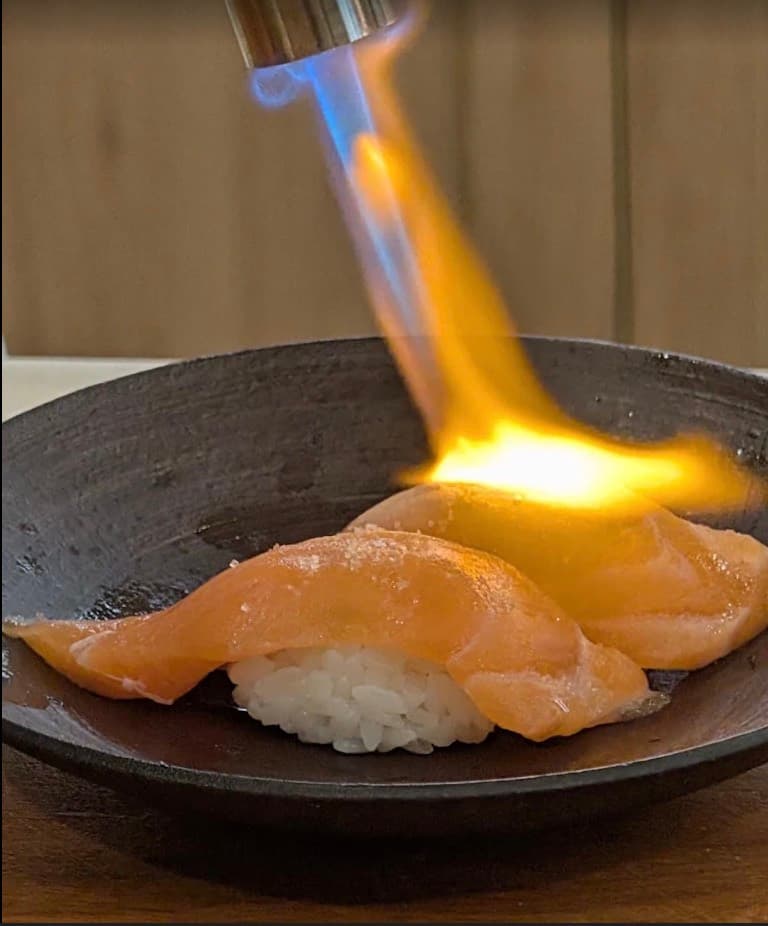 Sushi Seki, hai miếng nigiri cá hồi đang được khò lửa trong một chiếc tô sẫm màu