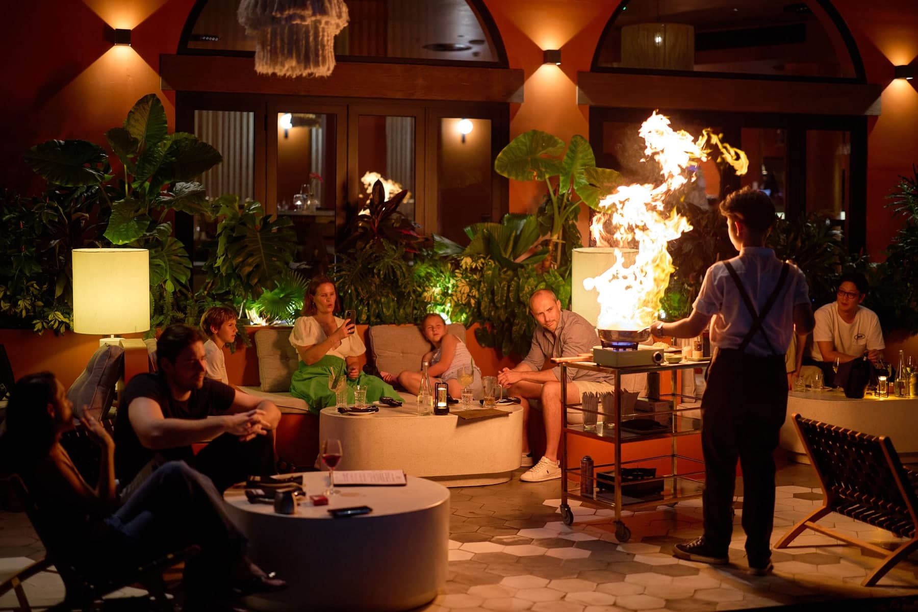 A pan flames on a trolley at The Gypsy Rooftop Restaurant & Bar while guests watch from low sofas among plants