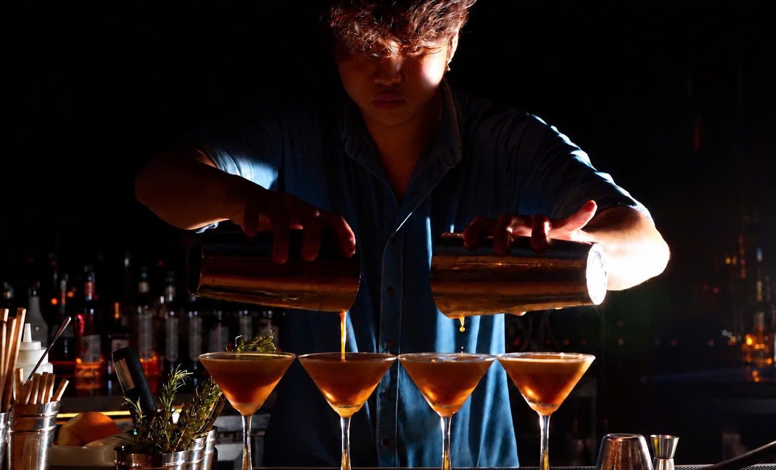 A bartender at The Gypsy Rooftop Restaurant & Bar pours from two tins into four cocktail glasses