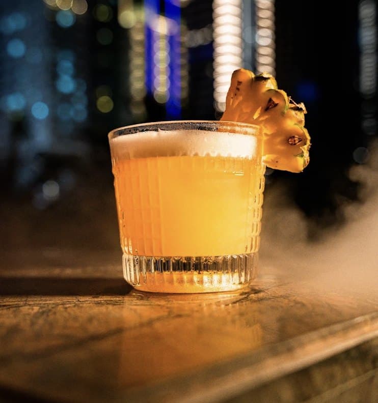 Một ly cocktail ở The Roof trong ly thủy tinh có vân, trang trí bằng miếng dứa, phía sau là ánh đèn thành phố