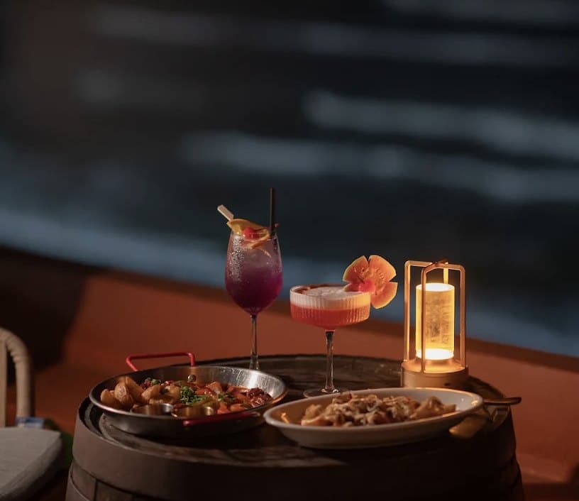 Hai ly cocktail, hai đĩa đồ ăn và một chiếc đèn nhỏ trên bàn thùng gỗ ở The Roof vào buổi tối