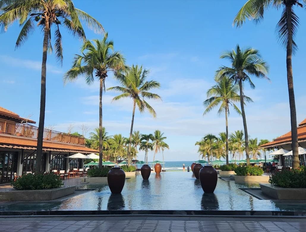 A long pool with dark jars and palm trees leading to the beach and the sea