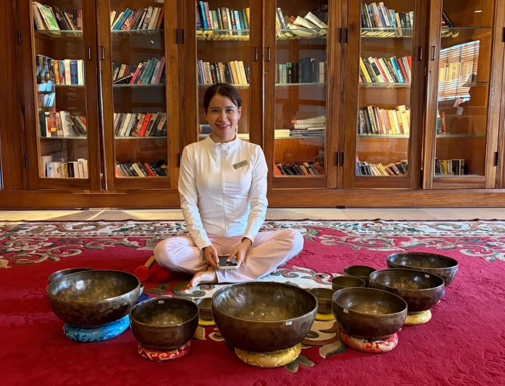 A woman in white sits on a red carpet among singing bowls at V-Senses Wellness & Spa