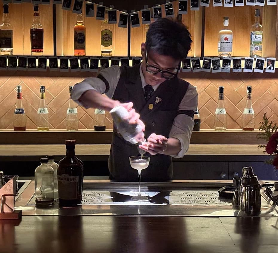 A bartender in a waistcoat straining a cocktail into a coupe behind the bar at Vấn Cocktail Bar in Da Nang