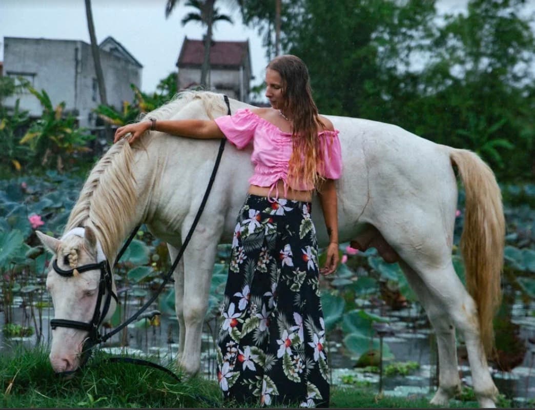 Vietnam Equestrian Club, a woman beside a white horse eating grass by a lotus pond, with houses behind