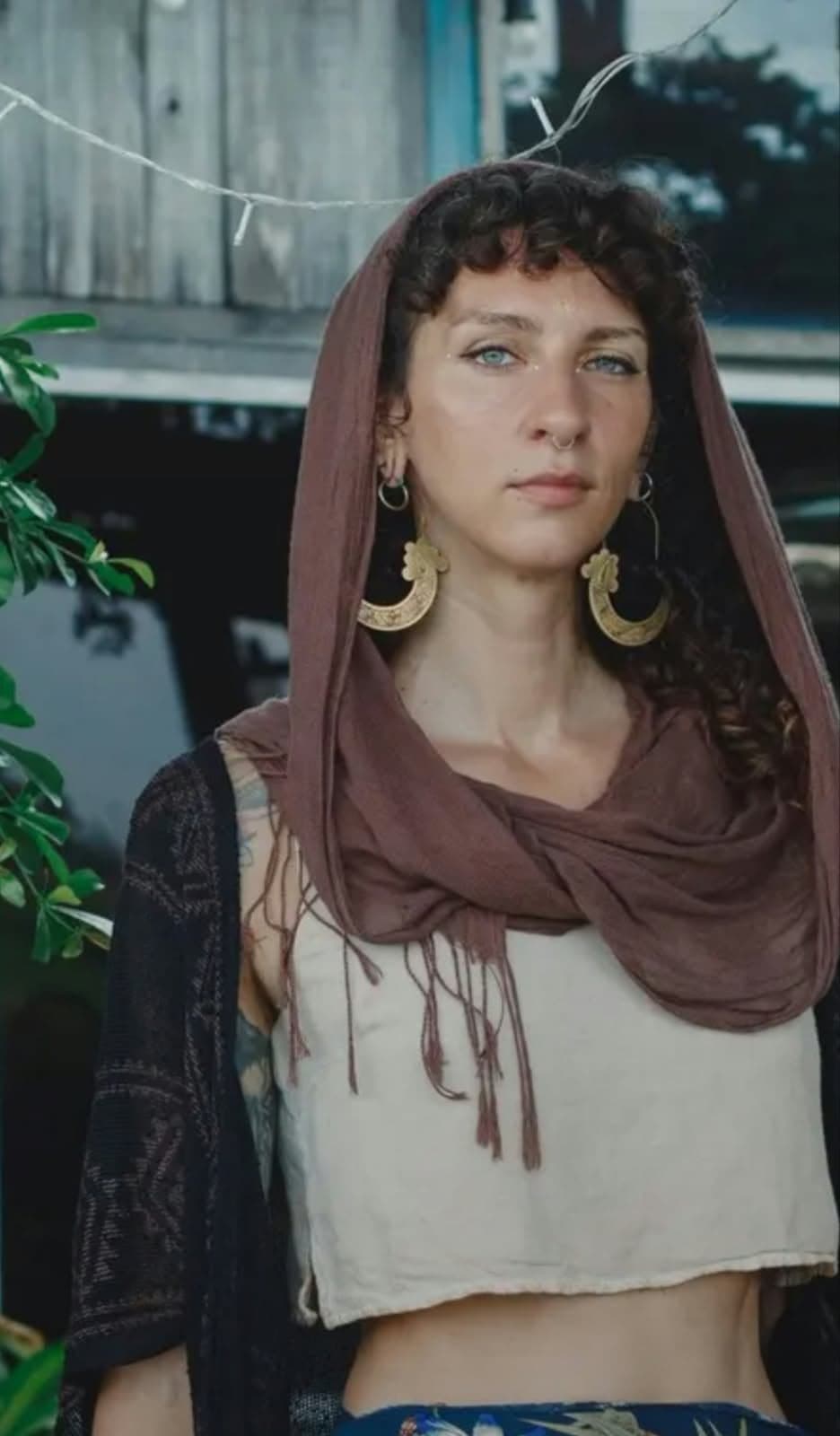 A woman in a brown hood and gold crescent earrings, standing by a wooden house with string lights